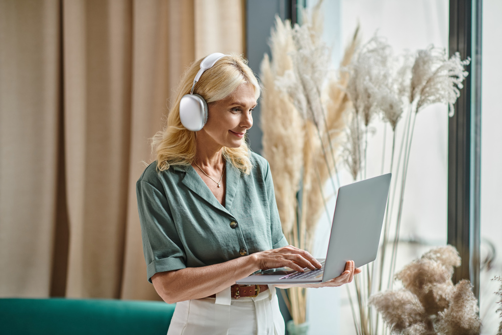 iloinen keski-ikäinen nainen langattomissa kuulokkeissa kuuntelee musiikkia ja käyttää kannettavaa tietokonetta kotona - Valokuva, kuva