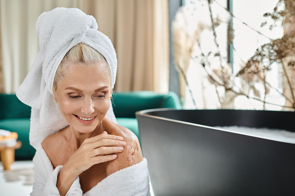 glückliche Frau mittleren Alters im weißen Bademantel und mit Handtuch auf dem Kopf, die Körperpeeling in der Nähe der Badewanne anwendet - Foto, Bild