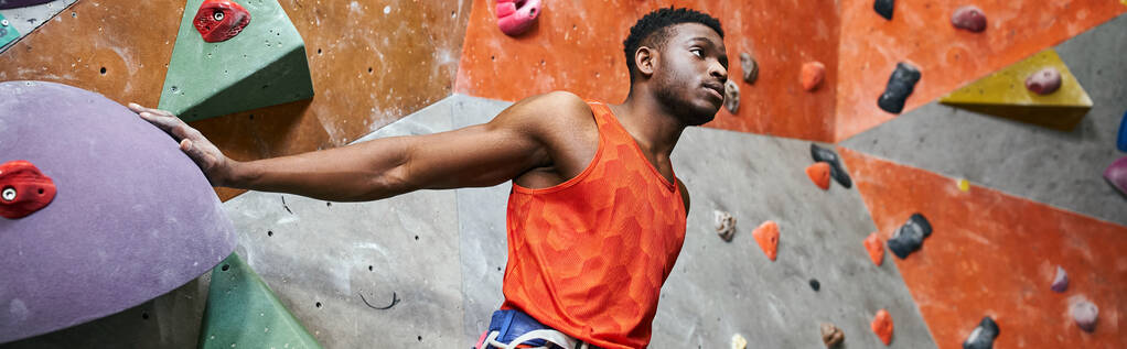 handsome african american man with alpine harness posing near climbing wall and looking away, banner - Photo, Image