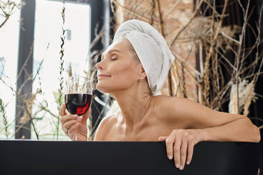 entspannte Frau mittleren Alters mit weißem Handtuch auf dem Kopf genießt den Geschmack von Rotwein beim Baden - Foto, Bild