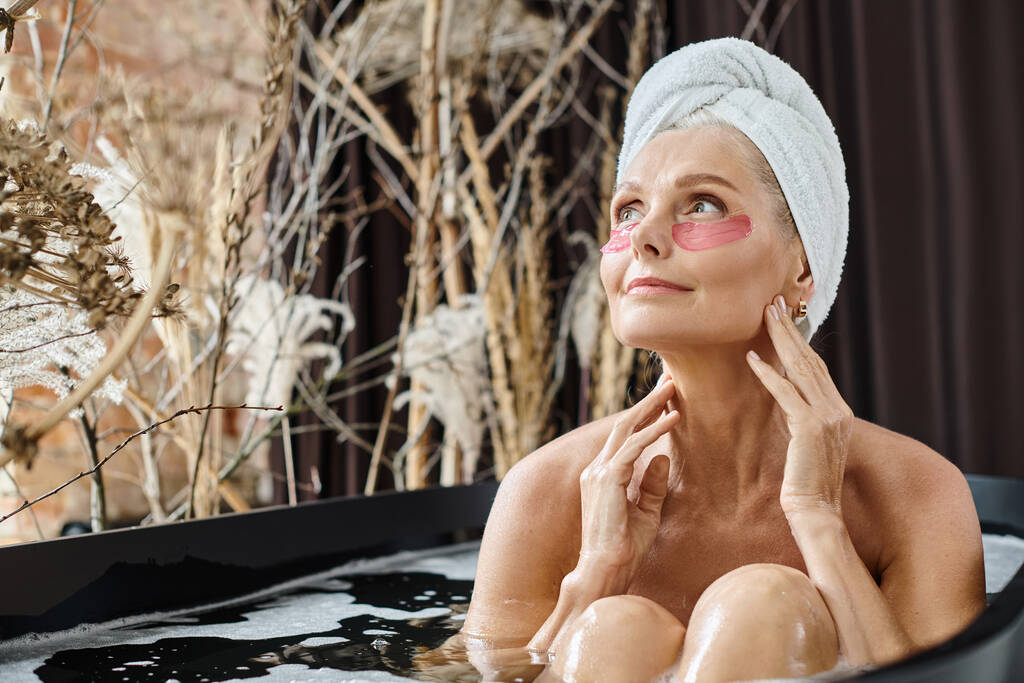 ontspannen vrouw van middelbare leeftijd met witte handdoek op het hoofd en onder de ogen pleisters nemen bad thuis - Foto, afbeelding