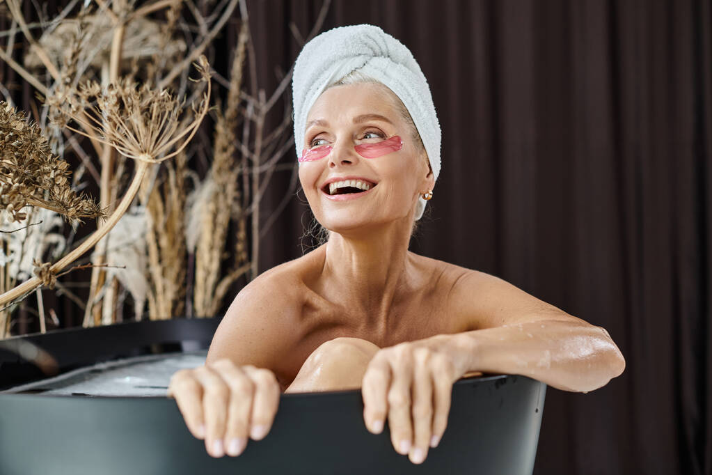 podekscytowana kobieta w średnim wieku z białym ręcznikiem na głowie i pod przepaskami na oczy biorąca kąpiel w domu - Zdjęcie, obraz