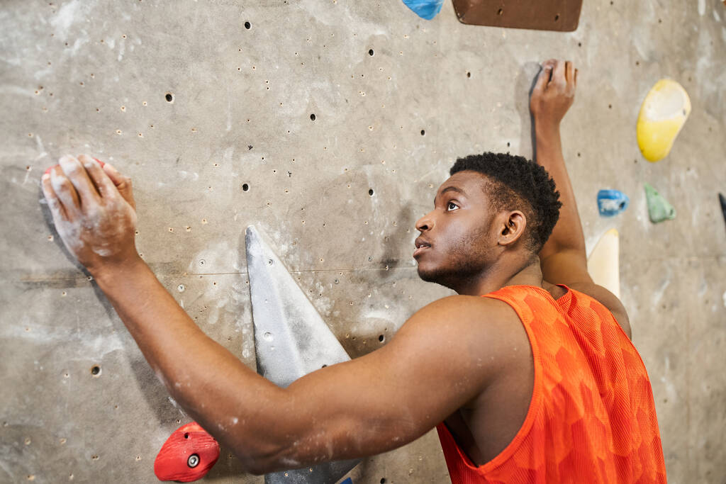 ハンサムな筋肉質のアフリカ系アメリカ人男性 オレンジ色のシャツで岩壁を登り,コンセプトをボルダリング - 写真・画像