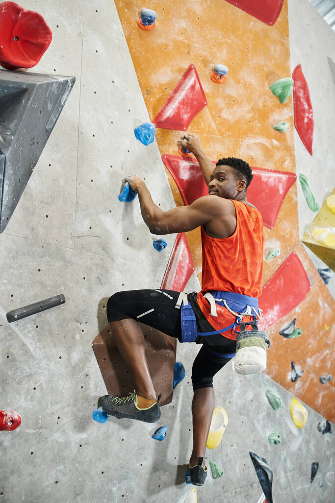 vertical shot of young african american man in orange shirt climbing up wall and looking ta camera - Photo, Image