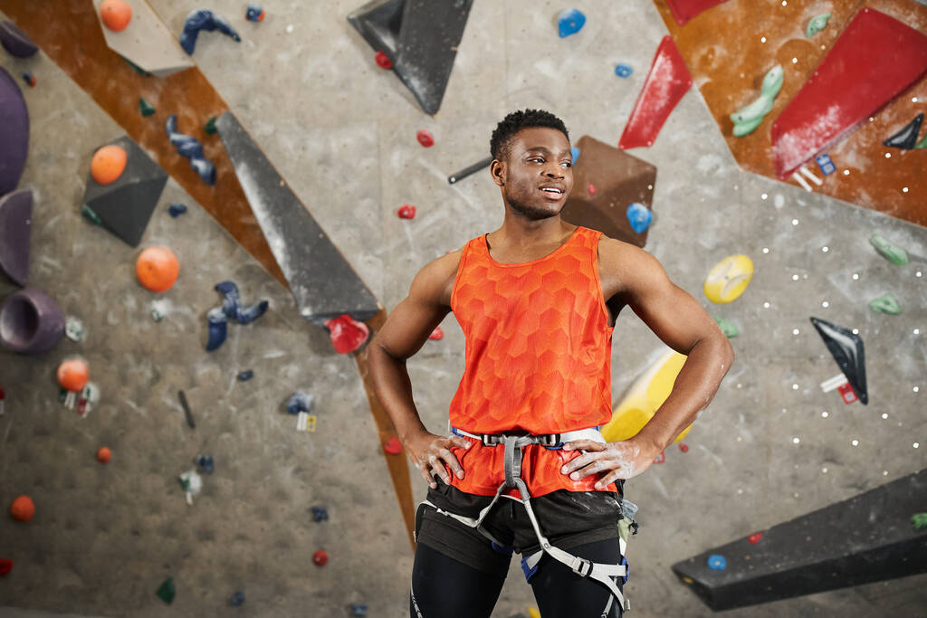 atlético hombre afroamericano en camisa naranja con arnés alpino posando con las manos en las caderas - Foto, Imagen