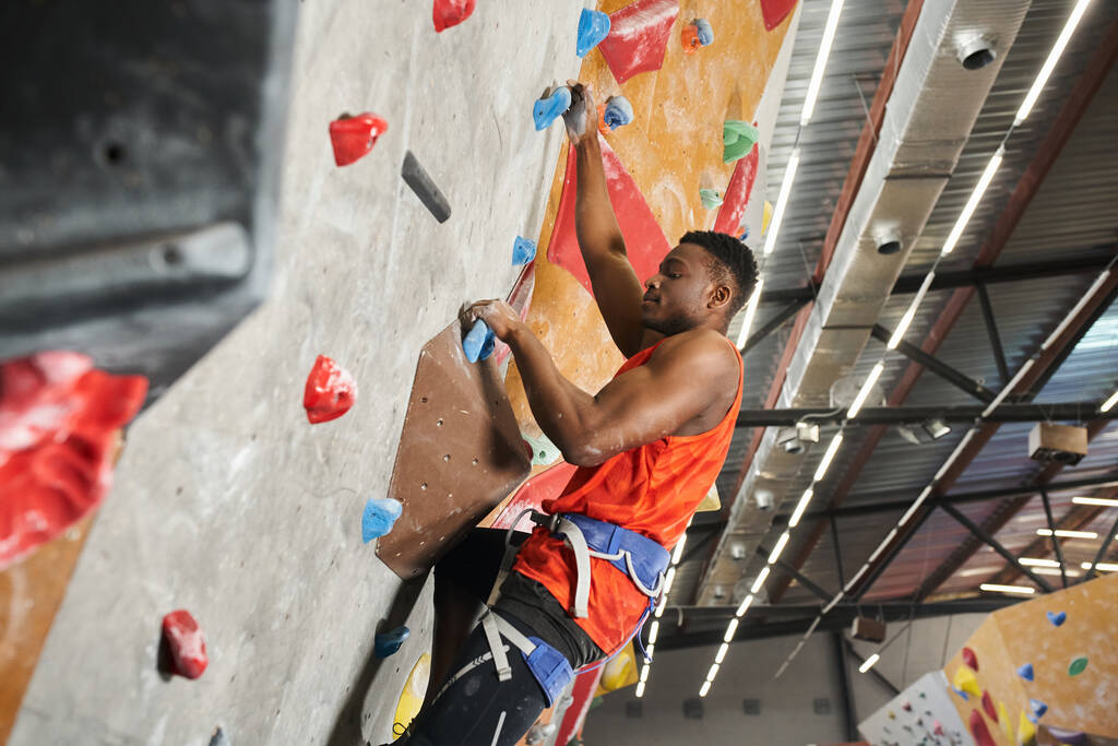 young african american man with alpine harness posing in profile while climbing up bouldering wall - Photo, Image