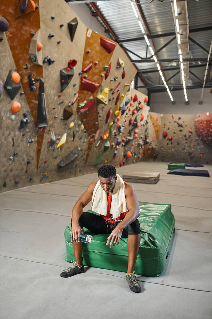 muscular african american man on crash pad holding water bottle with bouldering wall on backdrop - Photo, Image