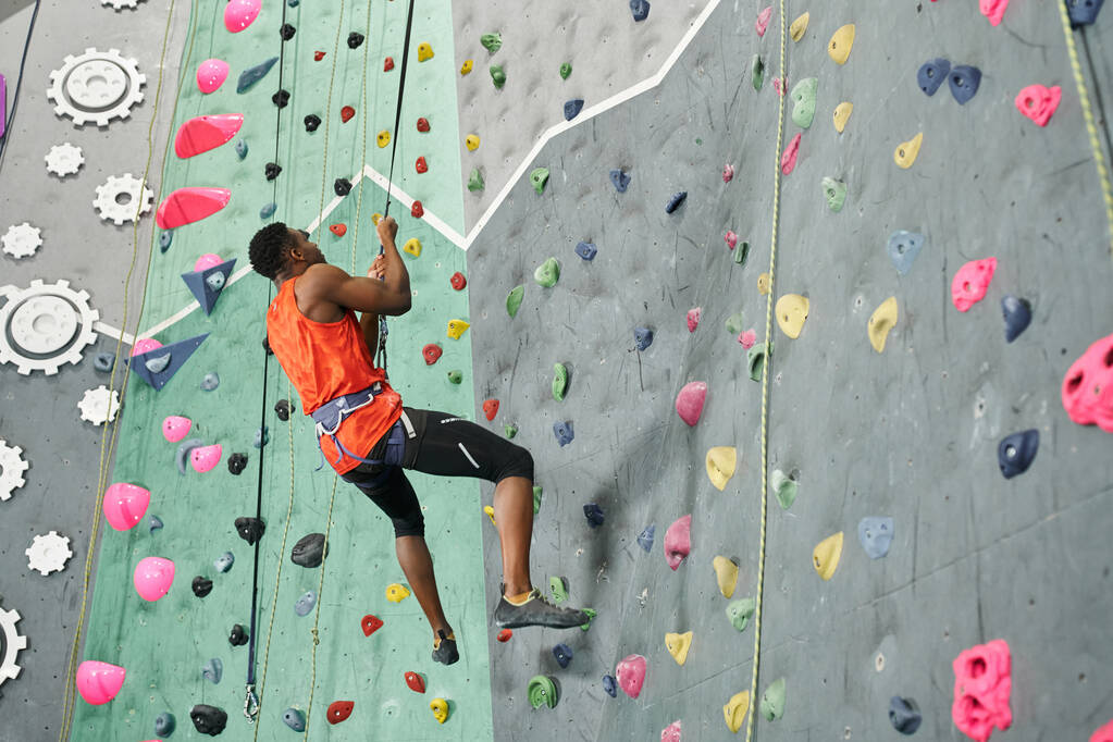 athletic african american man in orange shirt hanging on his climbing rope and looking up, rock wall - Photo, Image