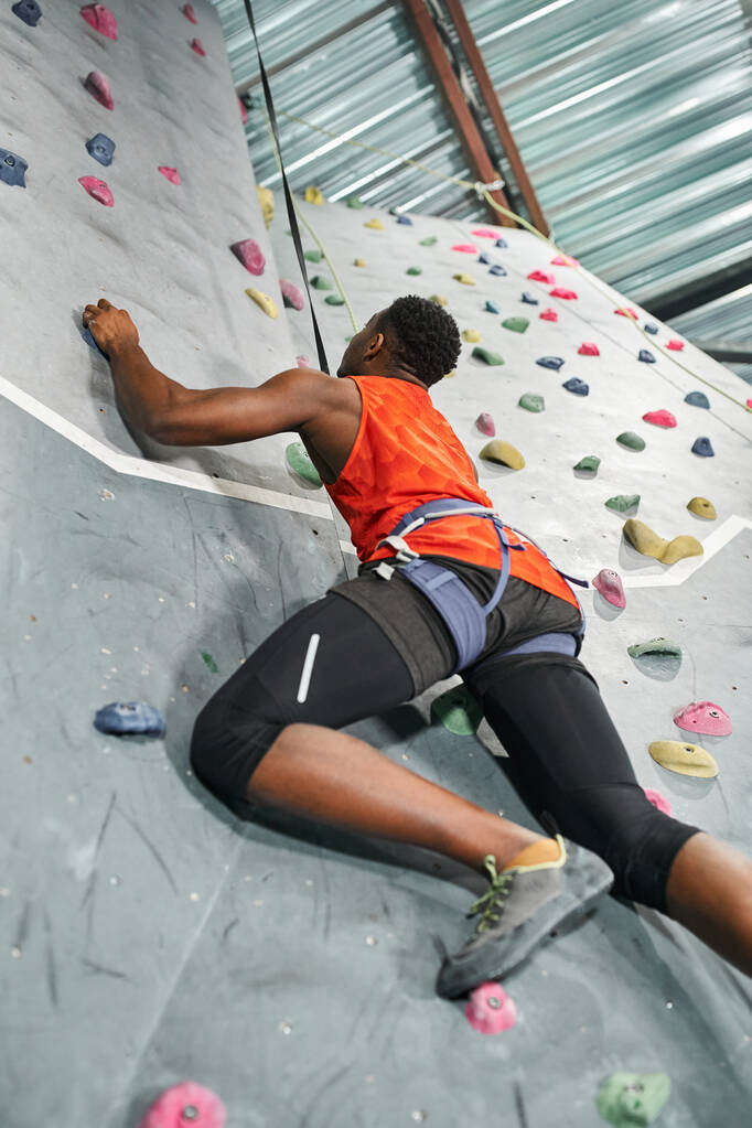 sportieve Afro-Amerikaanse man in oranje shirt met alpiene harnas klimmende rotswand, veiligheidstouw - Foto, afbeelding