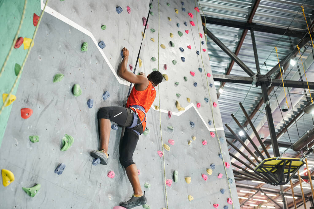 安全ロープとアルペンハーネスを使用して壁を登るオレンジ色のシャツの強いアフリカ系アメリカ人男性 - 写真・画像