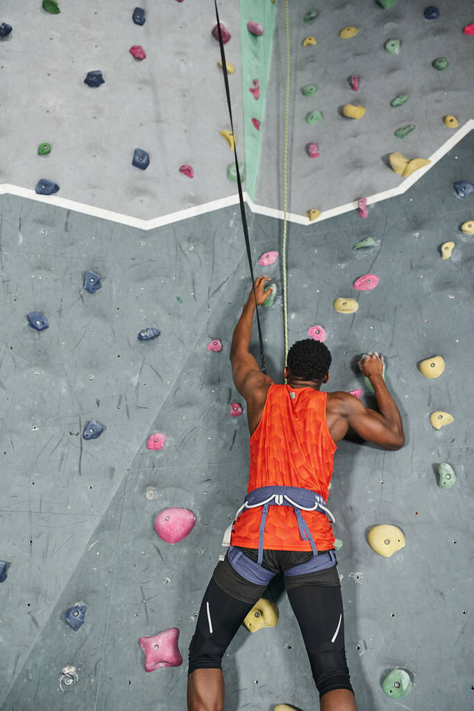 sterke jonge Afrikaan amerikaanse man met alpine harnas en veiligheid touw opstijgen muur, bouldering - Foto, afbeelding
