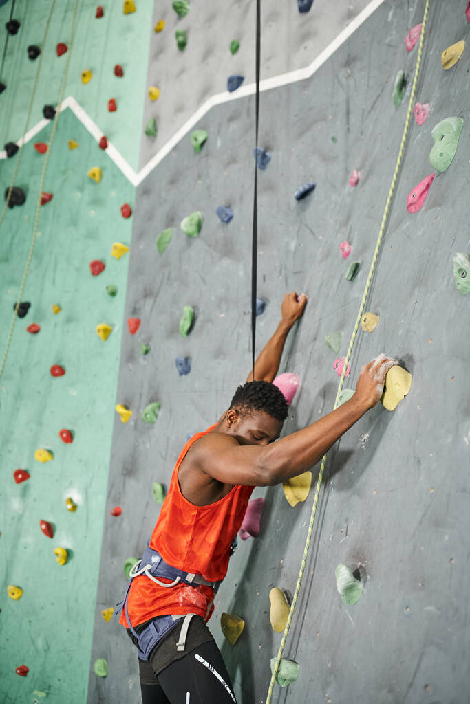 ハンサムなアフリカ系アメリカ人男性 オレンジ色のシャツ アルペンハーネスが壁を登る - 写真・画像