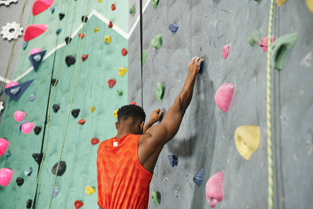 vista trasera del hombre afroamericano en camisa naranja trepando por la pared de bouldering con cuerda de seguridad - Foto, imagen