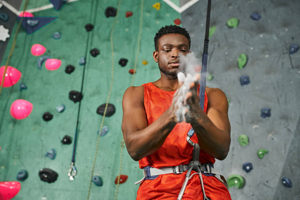 ロックウォールを登る前にジムチョークを使用してオレンジ色のシャツのハンサムな運動アフリカ系アメリカ人男性 - 写真・画像