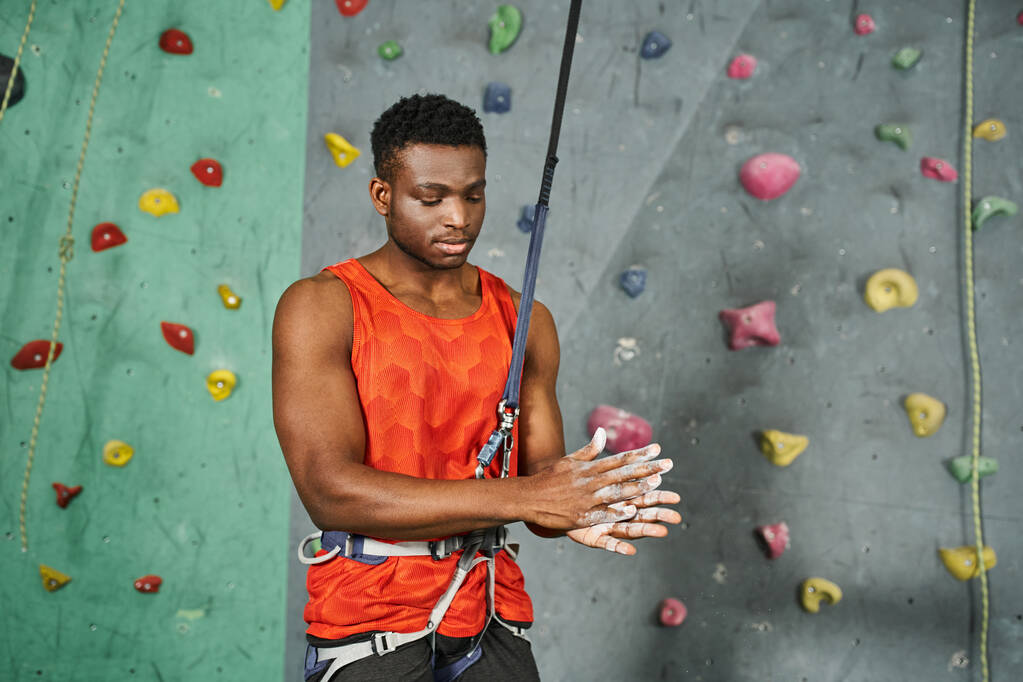 young athletic african american man in orange shirt with safety rope using talc powder, bouldering - Photo, Image