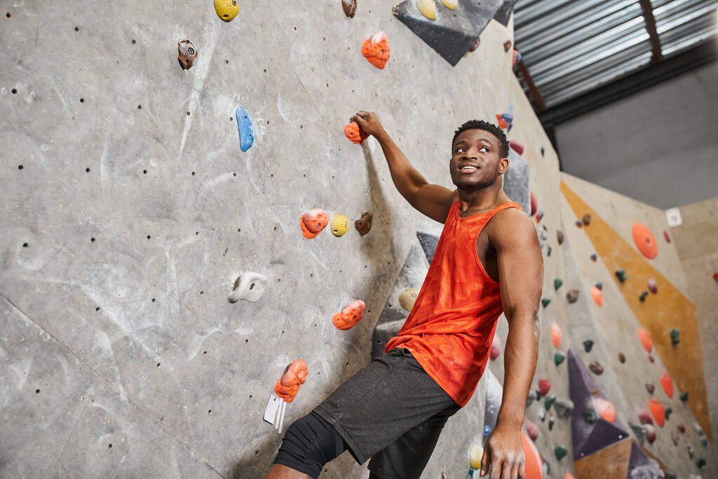 cheerful african american man in orange shirt ascending up rock wall and smiley looking away - Photo, Image