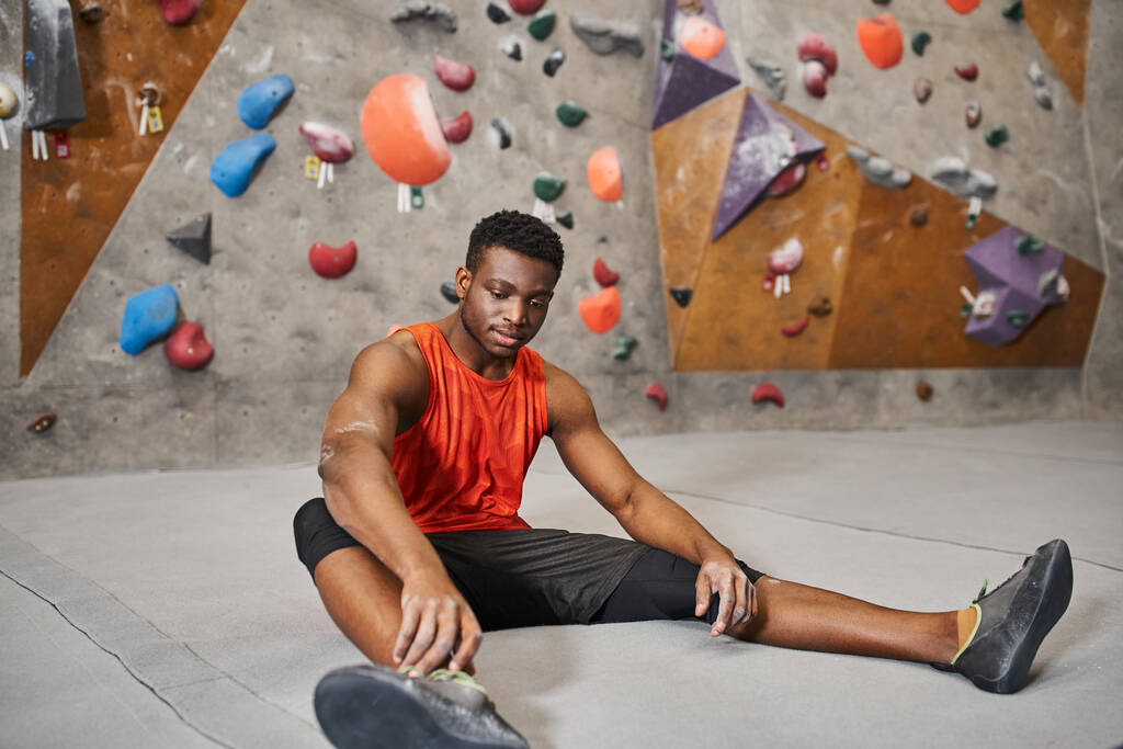 guapo joven afroamericano hombre de camisa naranja relajante en el suelo junto a la pared de bouldering - Foto, Imagen