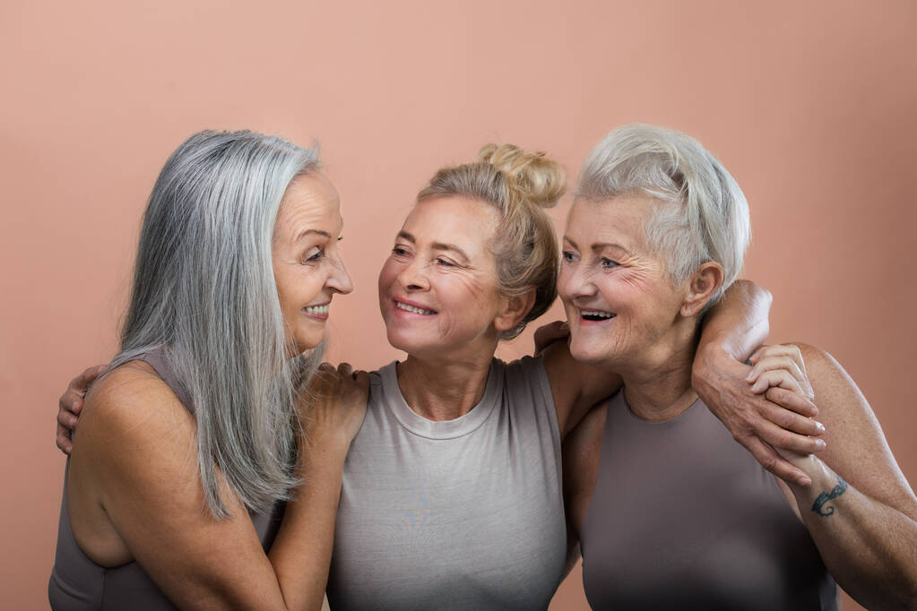 Studio portret trzech pięknych starszych kobiet z siwymi włosami. Starsze przyjaciółki trzymają się i uśmiechają. Odizolowany na brązowym różowym tle. Przestrzeń kopiowania. - Zdjęcie, obraz