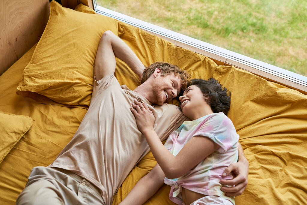 вид зверху щасливий чоловік обіймає азіатську дівчину і лежить разом на жовтому постілі вдома, пара - Фото, зображення