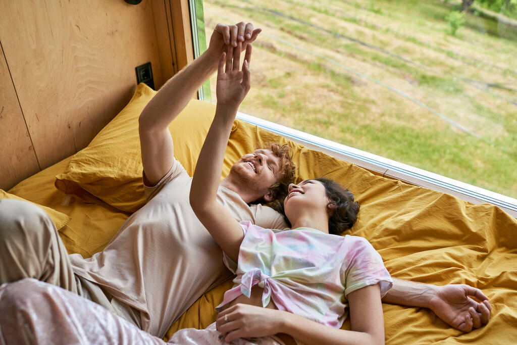 feliz hombre y joven asiático mujer tocando manos y acostado juntos en amarillo ropa de cama al lado de ventana - Foto, Imagen