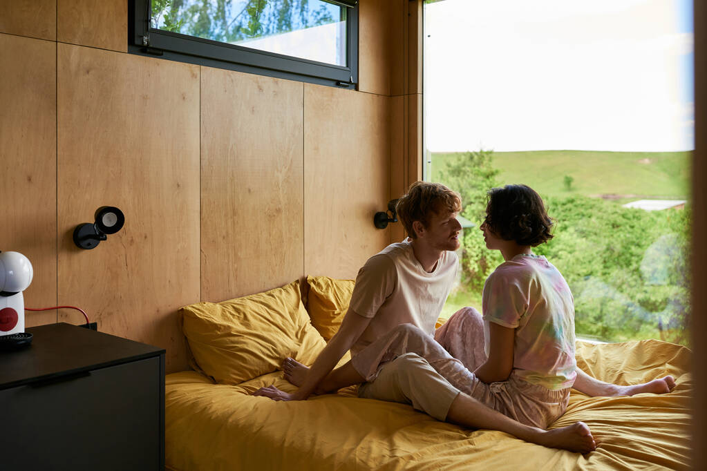 multiethnic couple looking at each other and sitting on bed in country house with forest view - Photo, Image