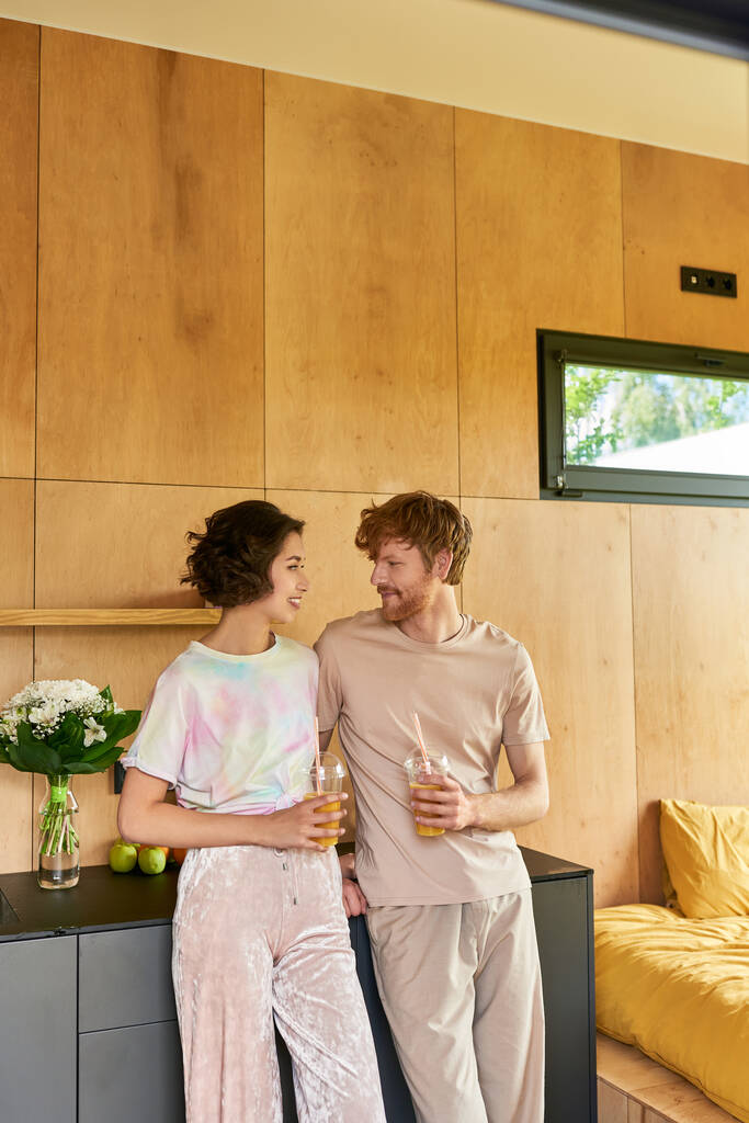 glückliches multikulturelles Paar, das morgens im Pyjama steht und Gläser mit Orangensaft in der Hand hält - Foto, Bild