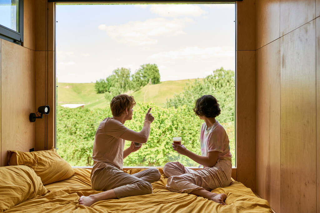 vista trasera del hombre apuntando a los árboles verdes detrás de la ventana, pareja sosteniendo tazas de café en el dormitorio - Foto, imagen