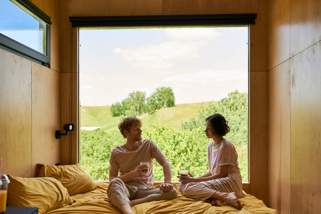 interracial couple holding cups of coffee and sitting on bed next to window with beautiful view - Photo, Image