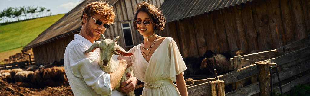 mosolygó többnemzetiségű pár esküvői ruhában és napszemüvegben ölelkező kecskék vidéken, transzparens - Fotó, kép