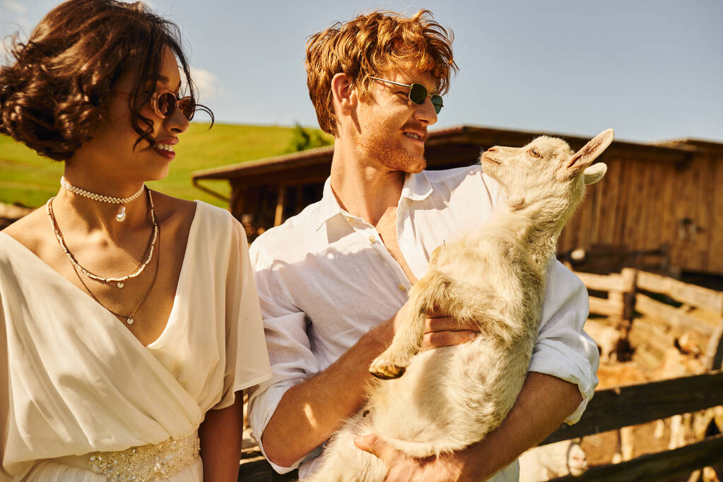 just married couple, redhead groom cuddling baby goat near asian bride in white dress, boho style - Photo, Image