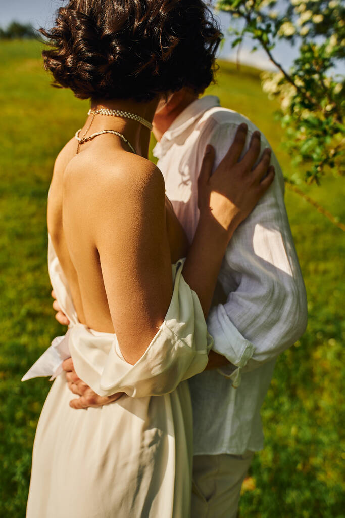 μελαχρινή νύφη σε λευκό φόρεμα που στέκεται με τον γαμπρό στον καταπράσινο κήπο, νιόπαντροι στην ύπαιθρο - Φωτογραφία, εικόνα