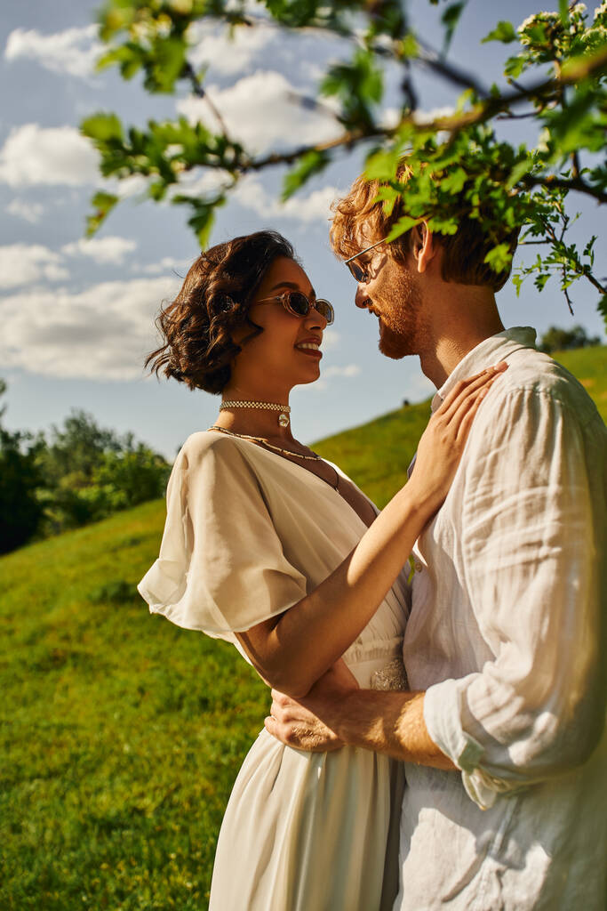 estilo boho, felices recién casados interracial en gafas de sol y vestido de novia abrazándose en el jardín verde - Foto, imagen