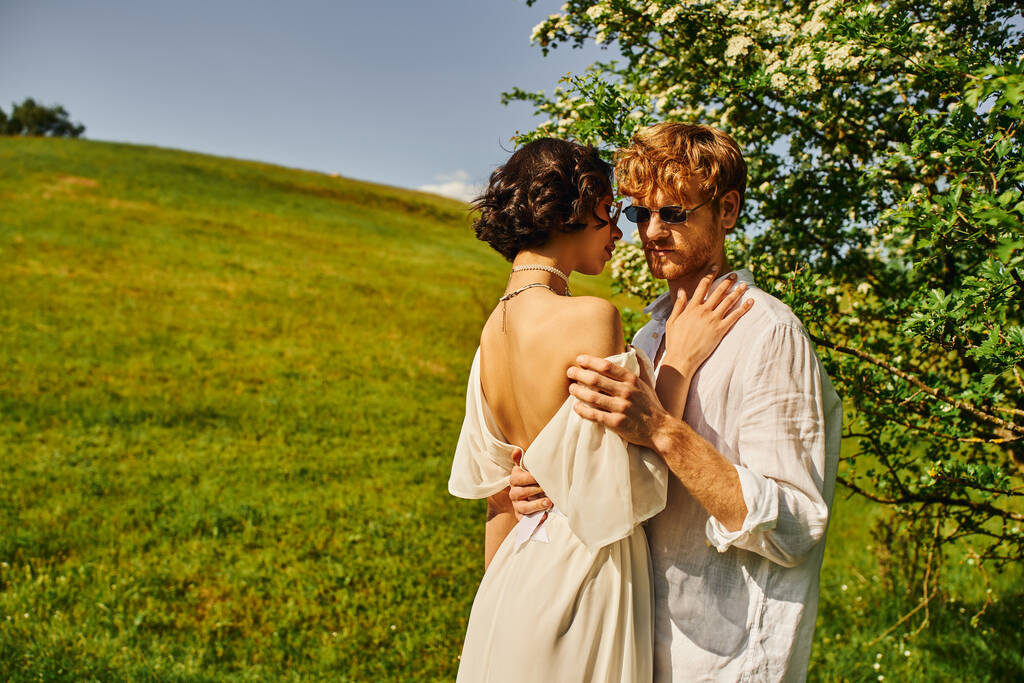 boho styl, szczęśliwy wielonarodowych nowożeńców w okularach przeciwsłonecznych i sukni ślubnej przytulanie na wsi - Zdjęcie, obraz