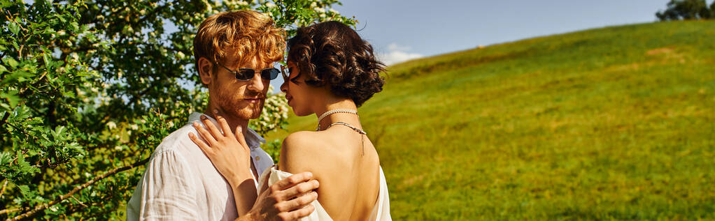 estilo boho, feliz pareja multiétnica en gafas de sol y vestido de novia abrazándose en el campo, bandera - Foto, Imagen