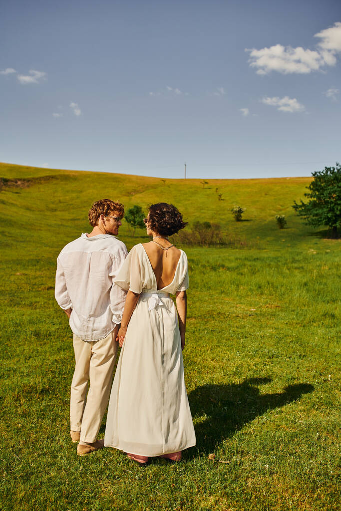 just married couple, asian bride in wedding dress holding hands with redhead groom in green field - Photo, Image