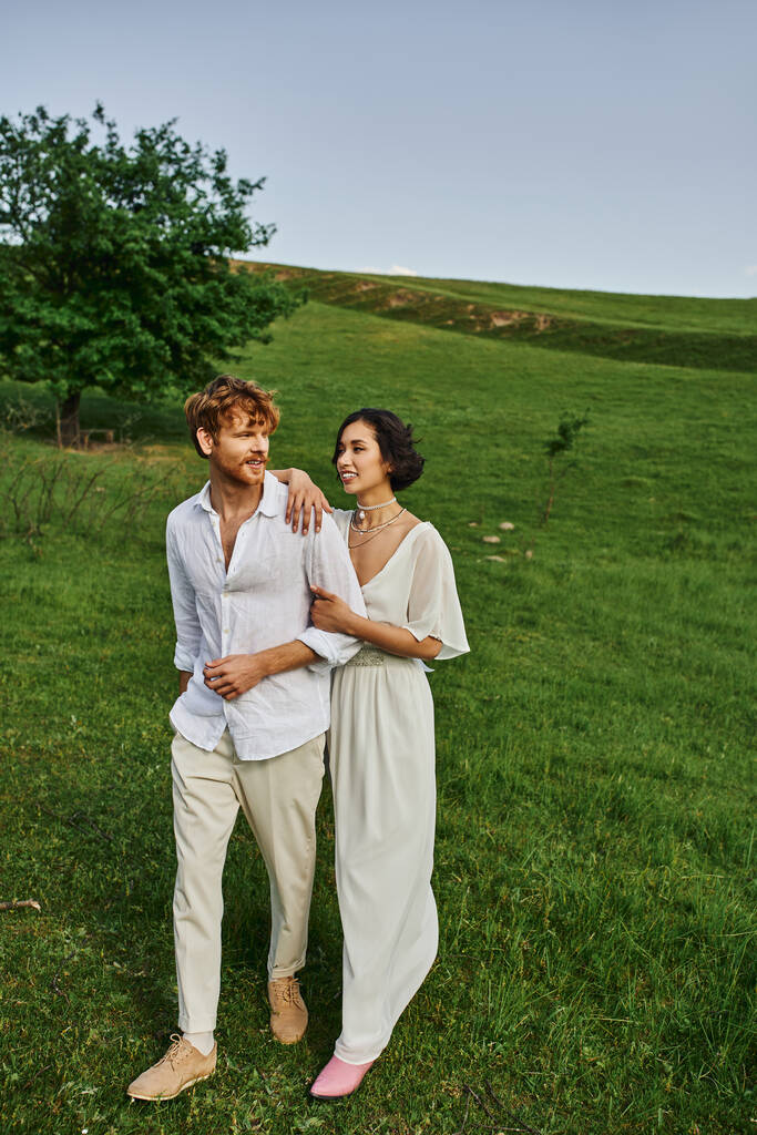 живописный пейзаж, счастливые молодожены в свадебном платье, гуляющие вместе в зеленом поле, только что поженились - Фото, изображение