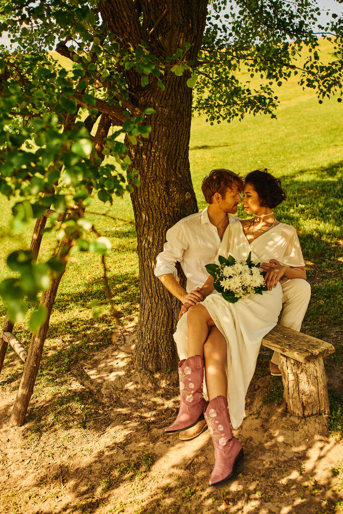 κοκκινομάλλα γαμπρός αγκαλιάζει ασιατική νύφη με μπουκέτο, ενώ κάθεται στον πάγκο κάτω από το δέντρο, ρουστίκ γάμο - Φωτογραφία, εικόνα