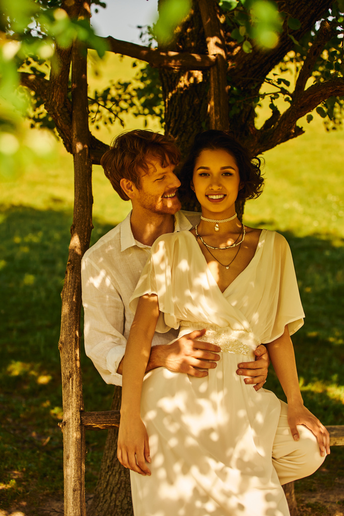 rustykalny ślub, uśmiechnięty rudy pan młody przytulający radosną azjatycką pannę młodą pod drzewem w słoneczny dzień - Zdjęcie, obraz