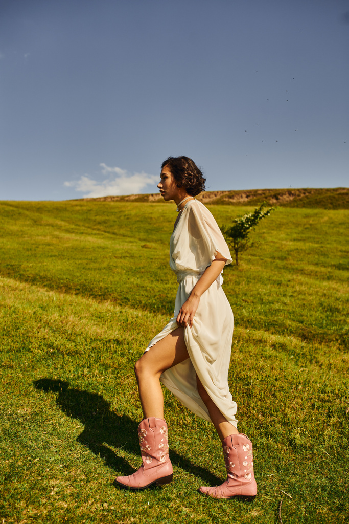vue latérale de jeune femme asiatique en robe de mariée et bottes de cow-boy marchant sur la prairie dans la campagne - Photo, image