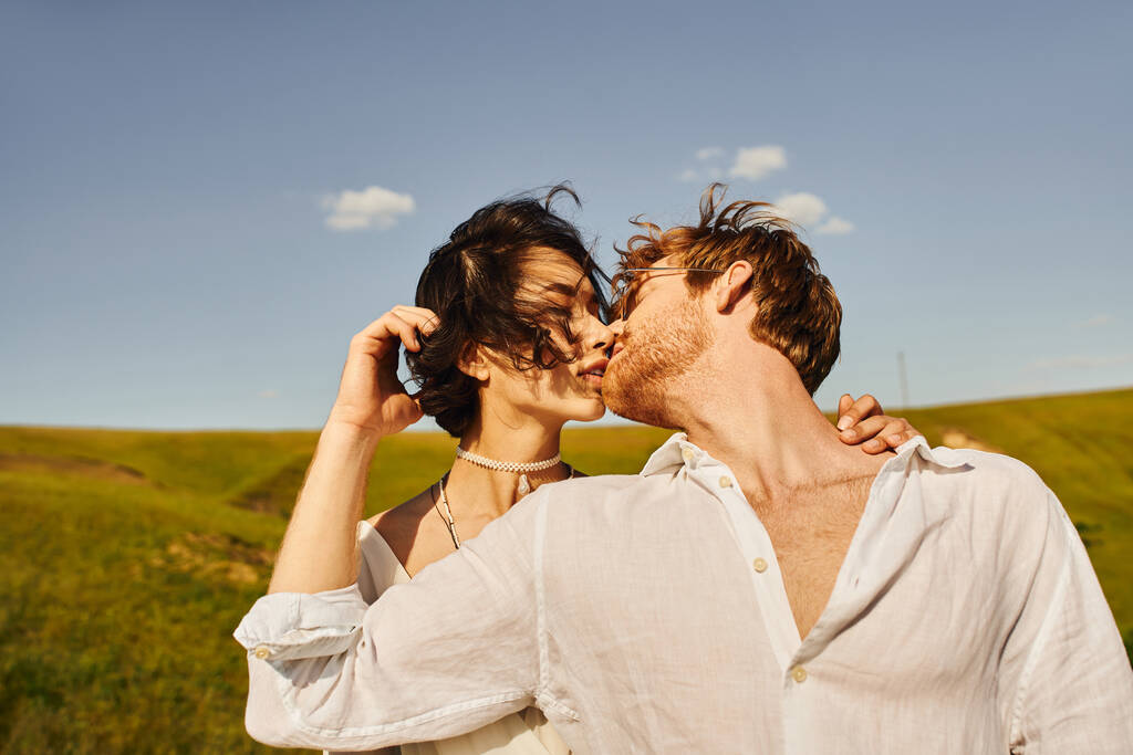 jen ženatý mezirasový pár líbání na větru pod modrým nebem, rustikální svatba v klidném prostředí - Fotografie, Obrázek