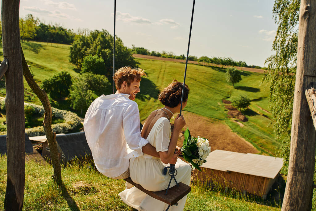 noivo ruiva alegre balançando com noiva em vestido branco no campo com paisagem pitoresca - Foto, Imagem