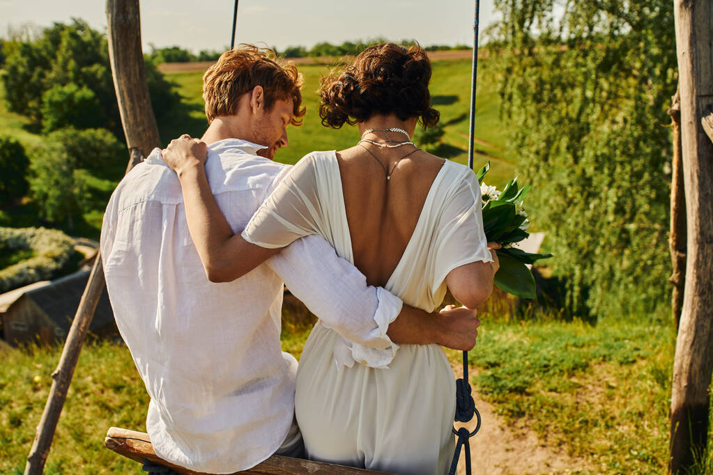 Вид сзади стильной молодоженов, катающихся на качелях в сельской местности, шумная свадьба - Фото, изображение