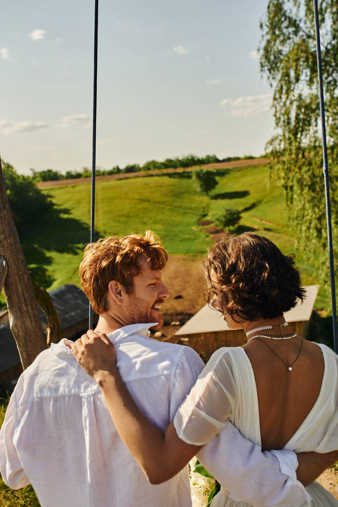 young interracial couple on swing in picturesque countryside setting, rural wedding - Photo, Image