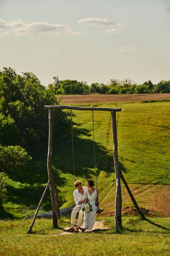 happy and stylish interracial newlyweds on swing in countryside with scenic landscape, tranquility - Photo, Image