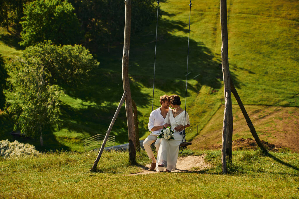 happy moment of just married multiethnic couple in boho style attire on swing in scenic countryside - Photo, Image