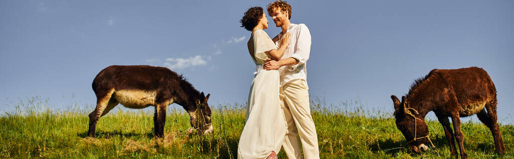 happy interracial newlyweds white attire embracing near donkeys grazing on meadow, banner - Photo, Image
