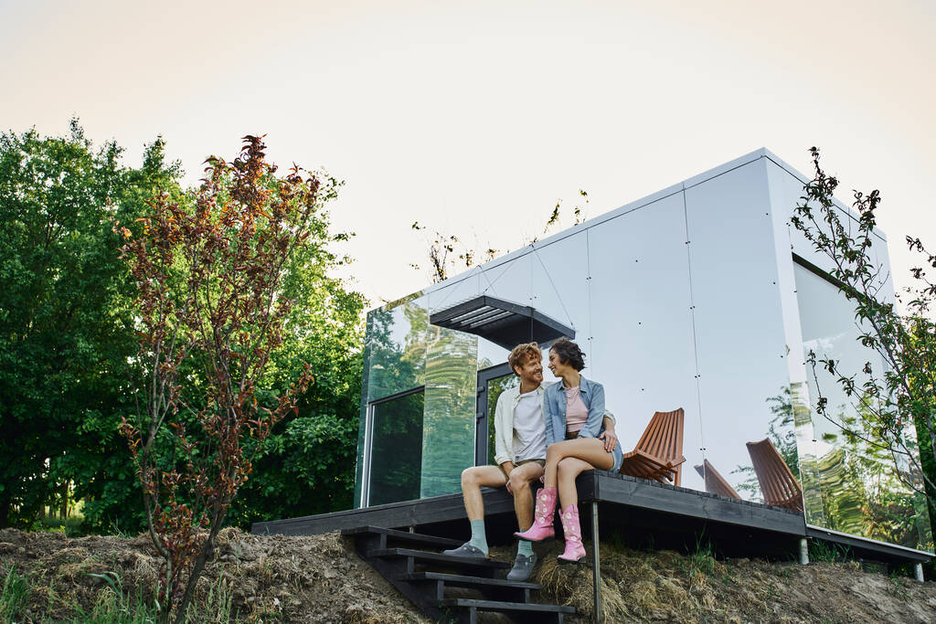 zorgeloze interraciale paar zitten op veranda van modern gras huis en glimlachen naar elkaar - Foto, afbeelding