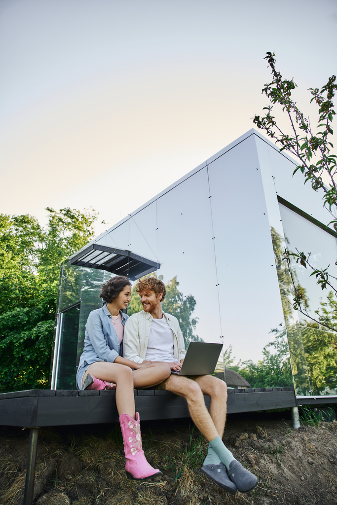щаслива міжрасова пара з ноутбуком, що сидить на ганку біля сучасного скляного будинку в сільській місцевості - Фото, зображення