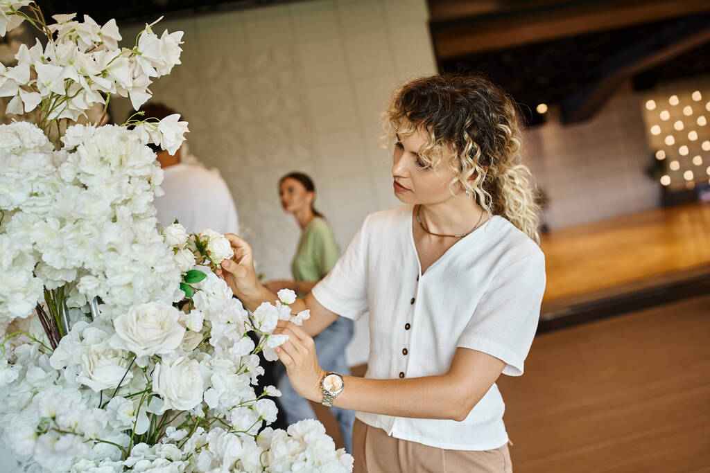 イベントホールのぼやけた背景にある花の装飾と同僚の近くのカメラを見ている楽しい女性 - 写真・画像