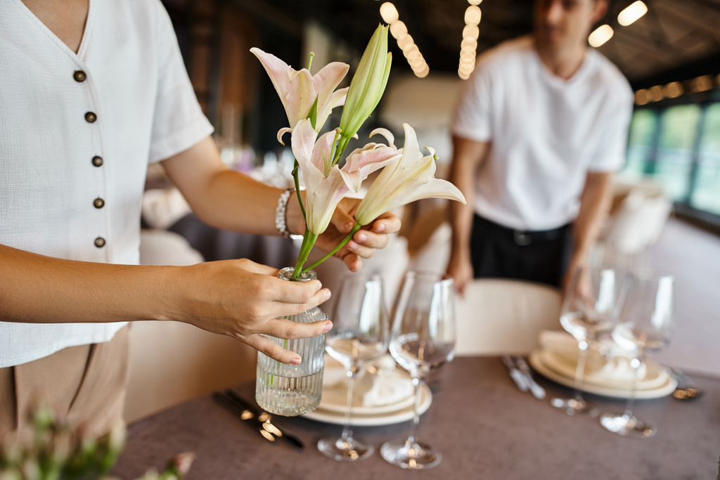 καλλιεργημένη άποψη του διακοσμητή κρατώντας βάζο και λουλούδια κοντά στο τραπέζι με εορταστική ρύθμιση στην αίθουσα εκδηλώσεων - Φωτογραφία, εικόνα